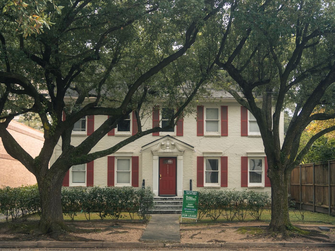 Brick two-storey house with red shutters under large oak trees
