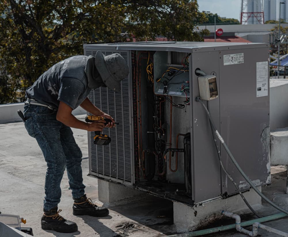Technician in a cap leans into an outdoor air conditioning unit on a rooftop to repair it